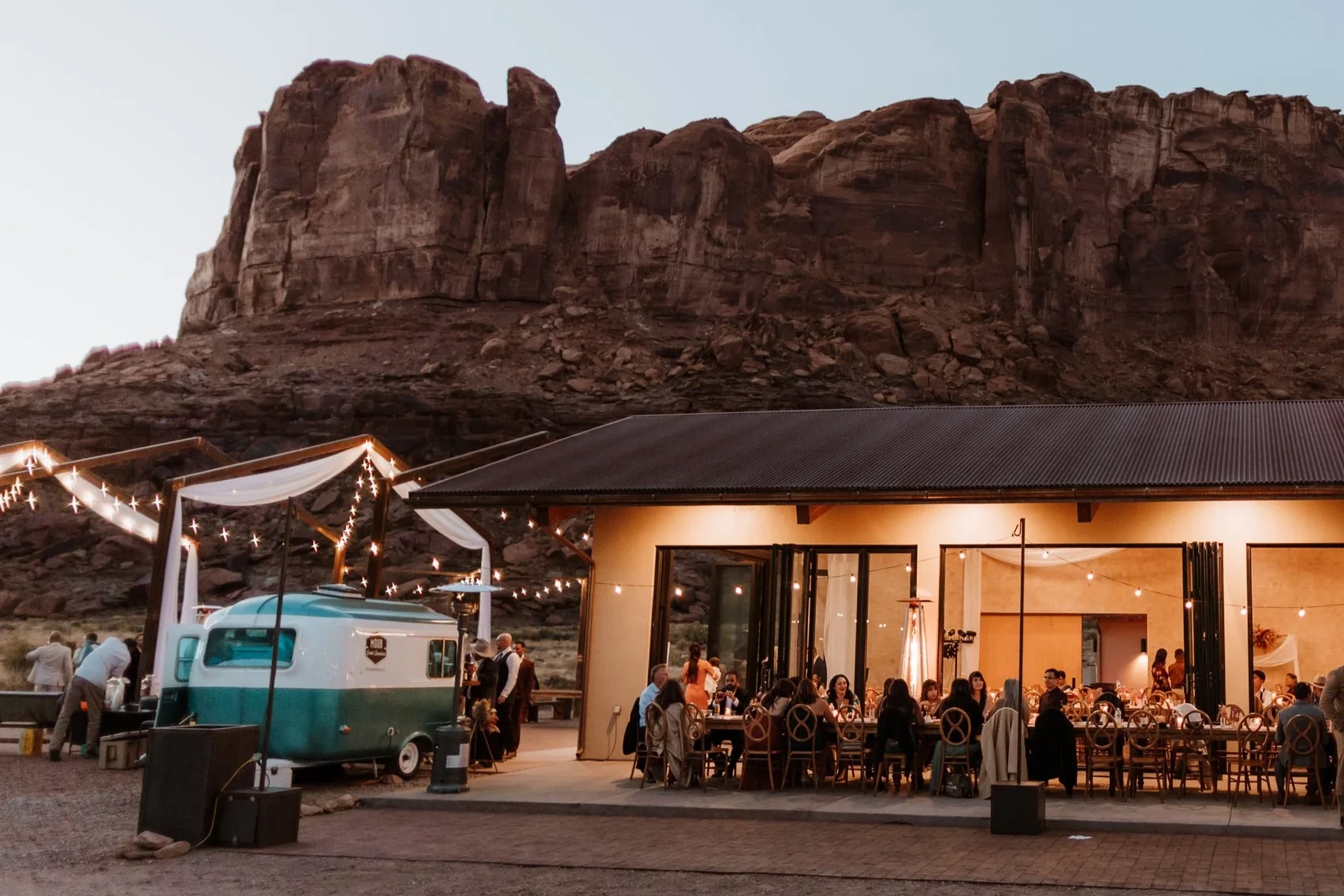 A gathering of people at an outdoor event with string lights, set against red rock formations at dusk, with a modern building and a vintage camper in the foreground.