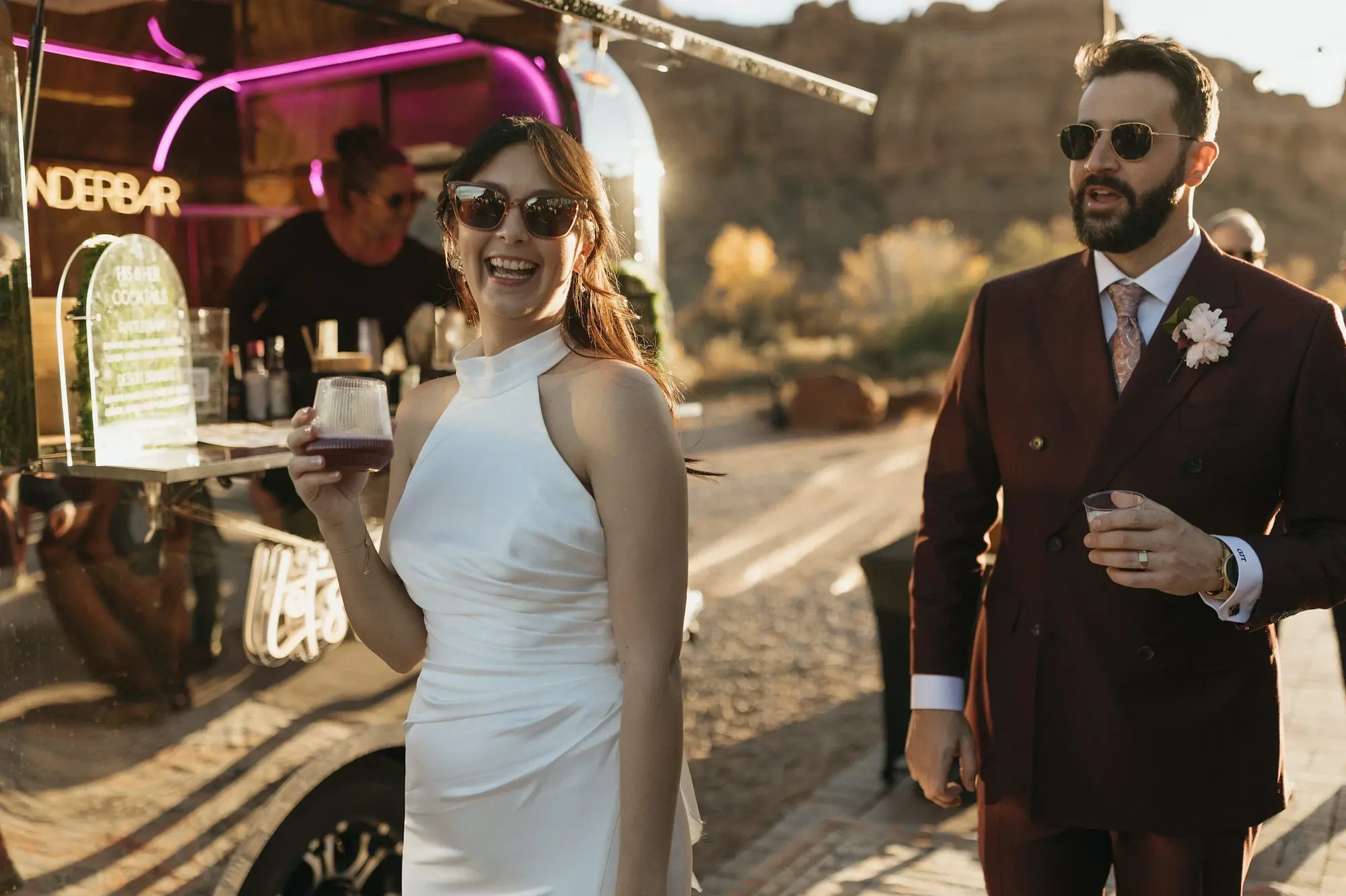 A woman in a white dress with large sunglasses is smiling and holding a drink, standing next to a man in a maroon suit with sunglasses, at an outdoor event during sunset.