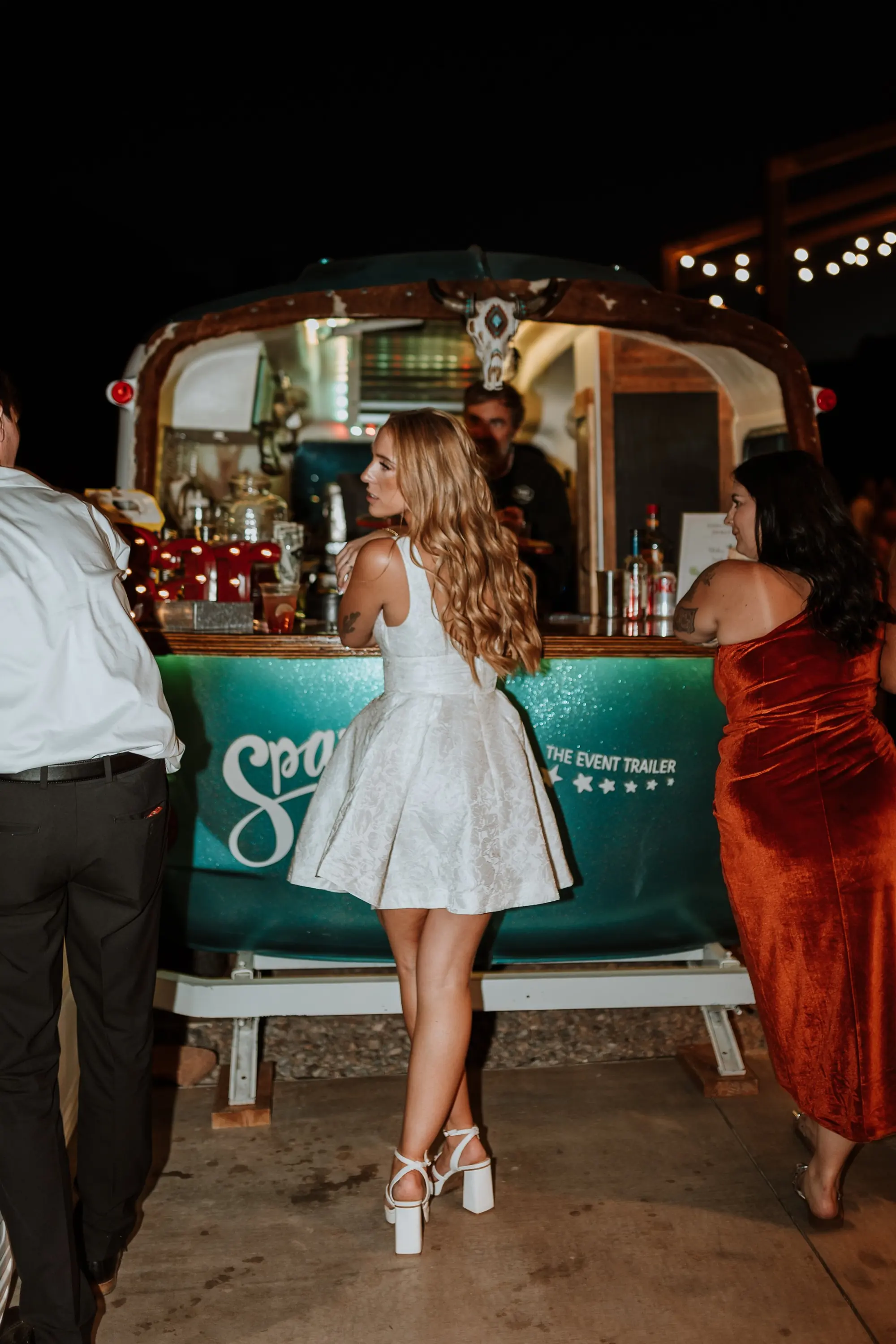 People gathered at a bar with a cowboy skull mounted on top, nighttime setting with string lights in the background.