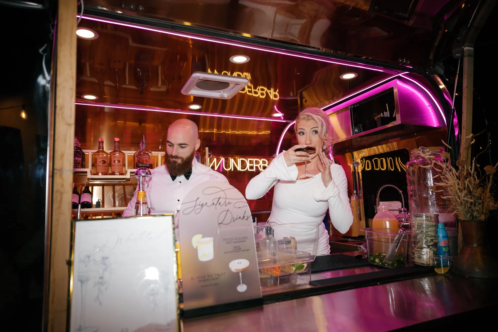 A woman with blonde and pink hair drinking from a cocktail glass behind a bar with bartender and bottles in the background, illuminated with pink neon lights.