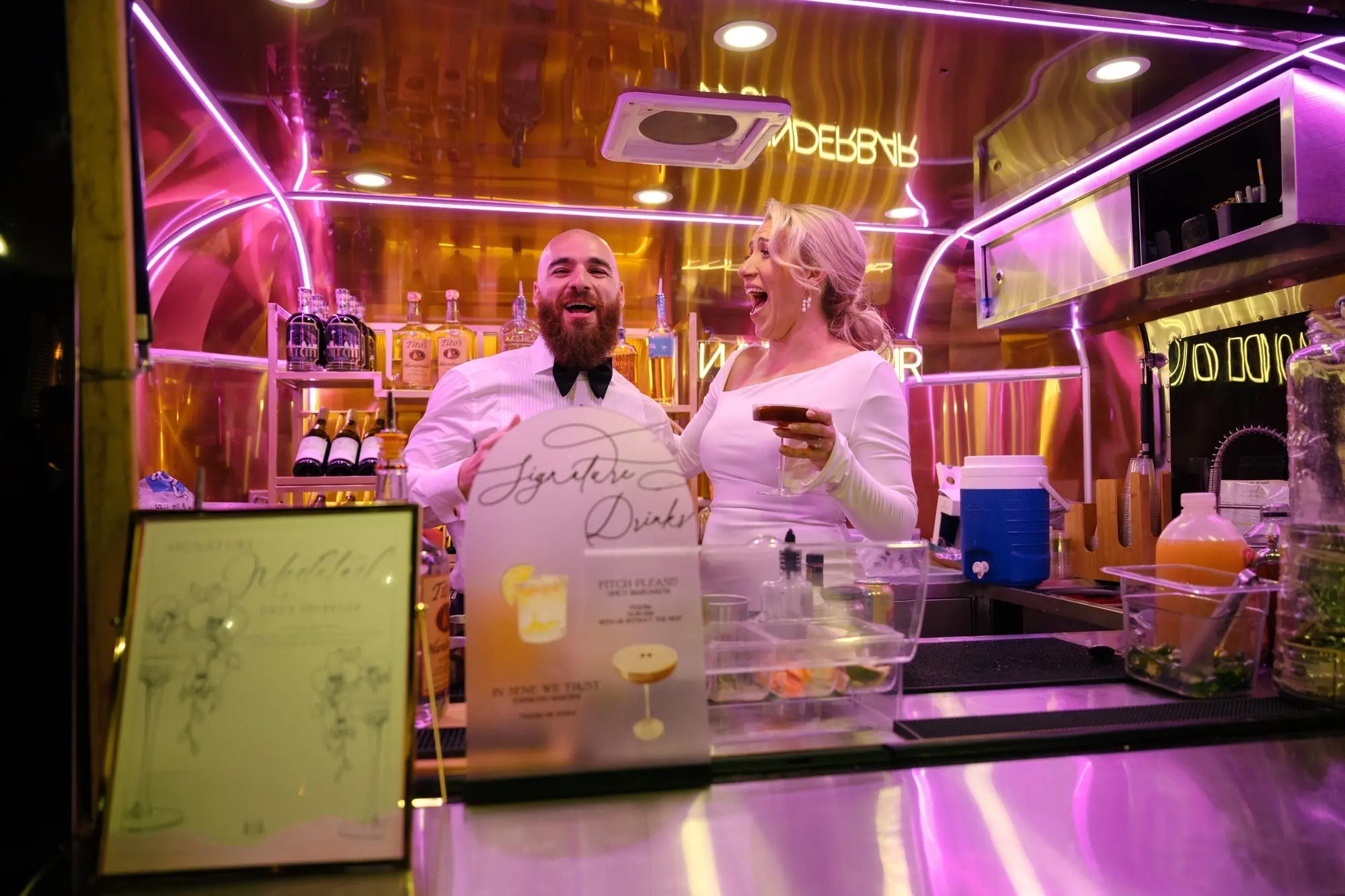 A man in a tuxedo with a bowtie and a woman in a white dress are smiling and laughing behind a bar counter illuminated with pink neon lights. The bar is stocked with liquor bottles and various bar equipment, and they seem to be enjoying their time to