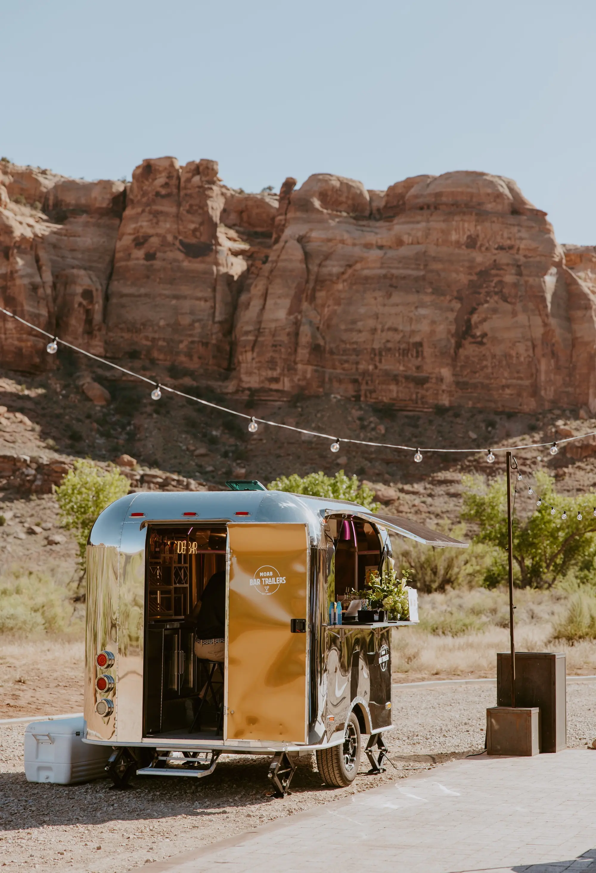 A shiny, metallic mobile bar trailer set up outdoors in a desert-like environment with large rock formations in the background. A string of lights is hung above the trailer, with a small table beside it and a food and drink sign on the side.