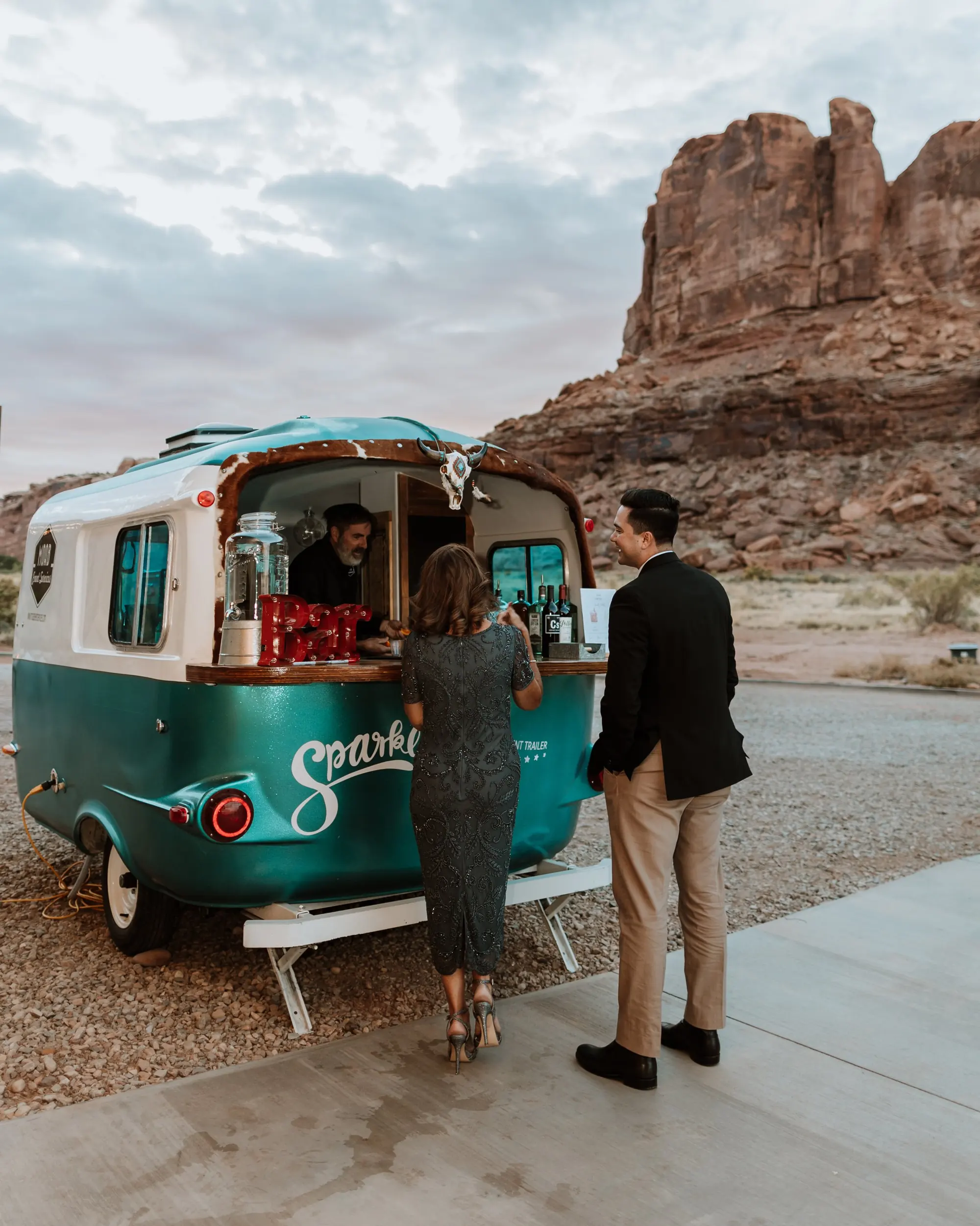 People at a food truck in a desert area with large rock formations and cloudy sky in the background.