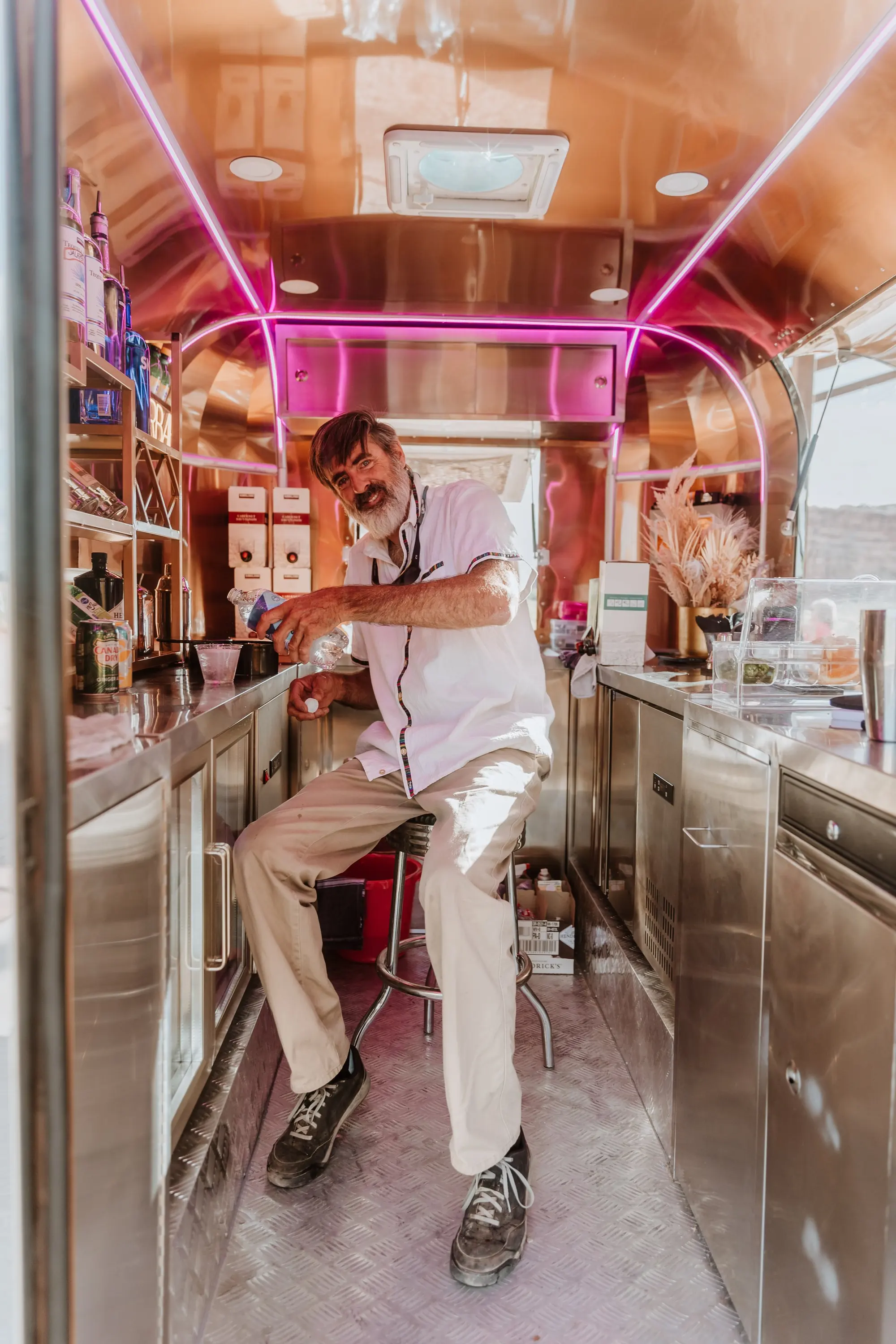 A man with grey hair and a beard sitting inside a small, shiny, copper-colored mobile bar or food truck, pouring a drink. He's wearing a white shirt, beige pants, and dark sneakers. The interior features stainless steel counters, shelves with bottles