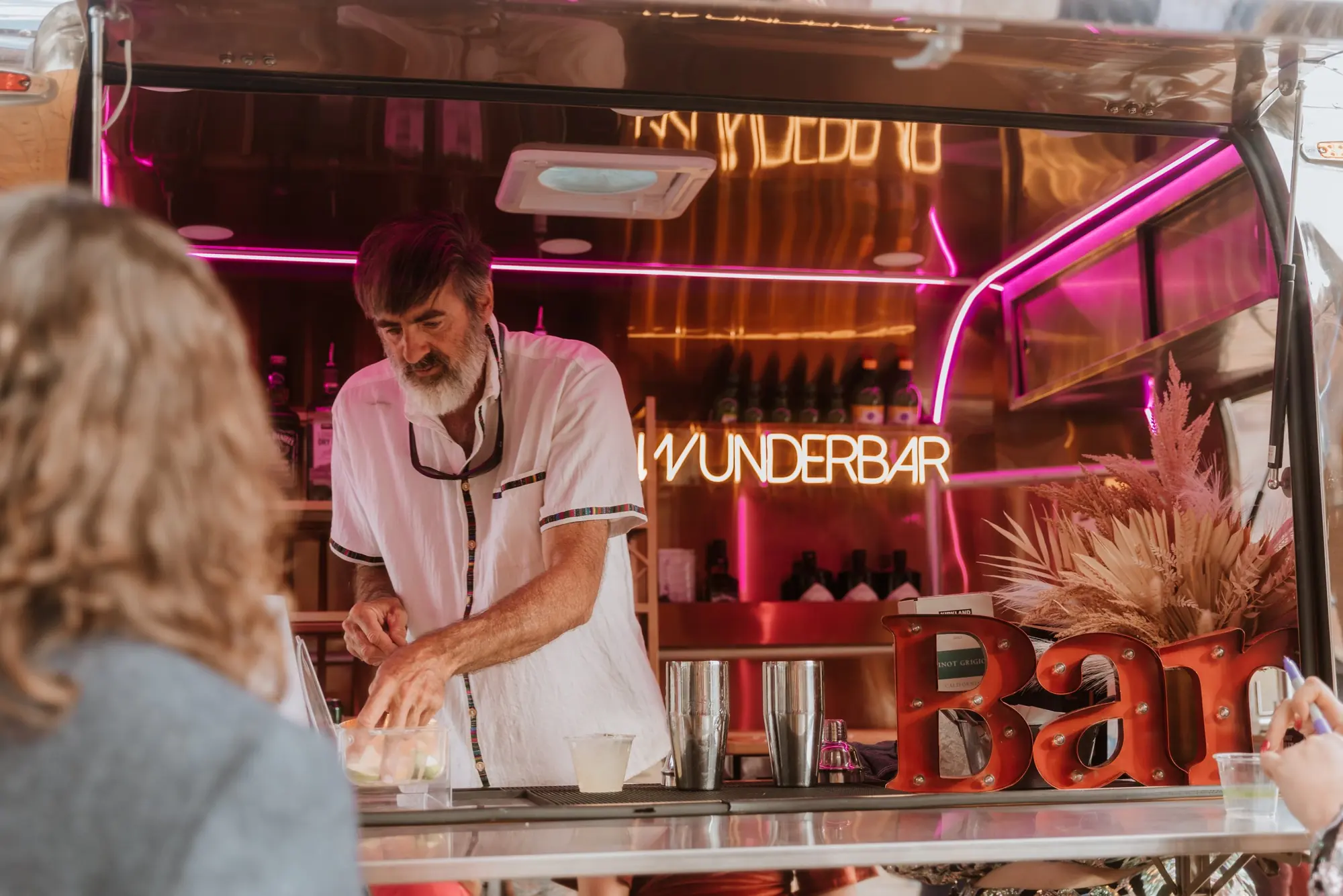 A man with dark hair and a beard working at a colorful, illuminated bar or food truck with a neon sign that reads 'UNDERBAR'. There are people in the foreground, including a blonde woman with curly hair and a child's hand holding a drink. The bar has
