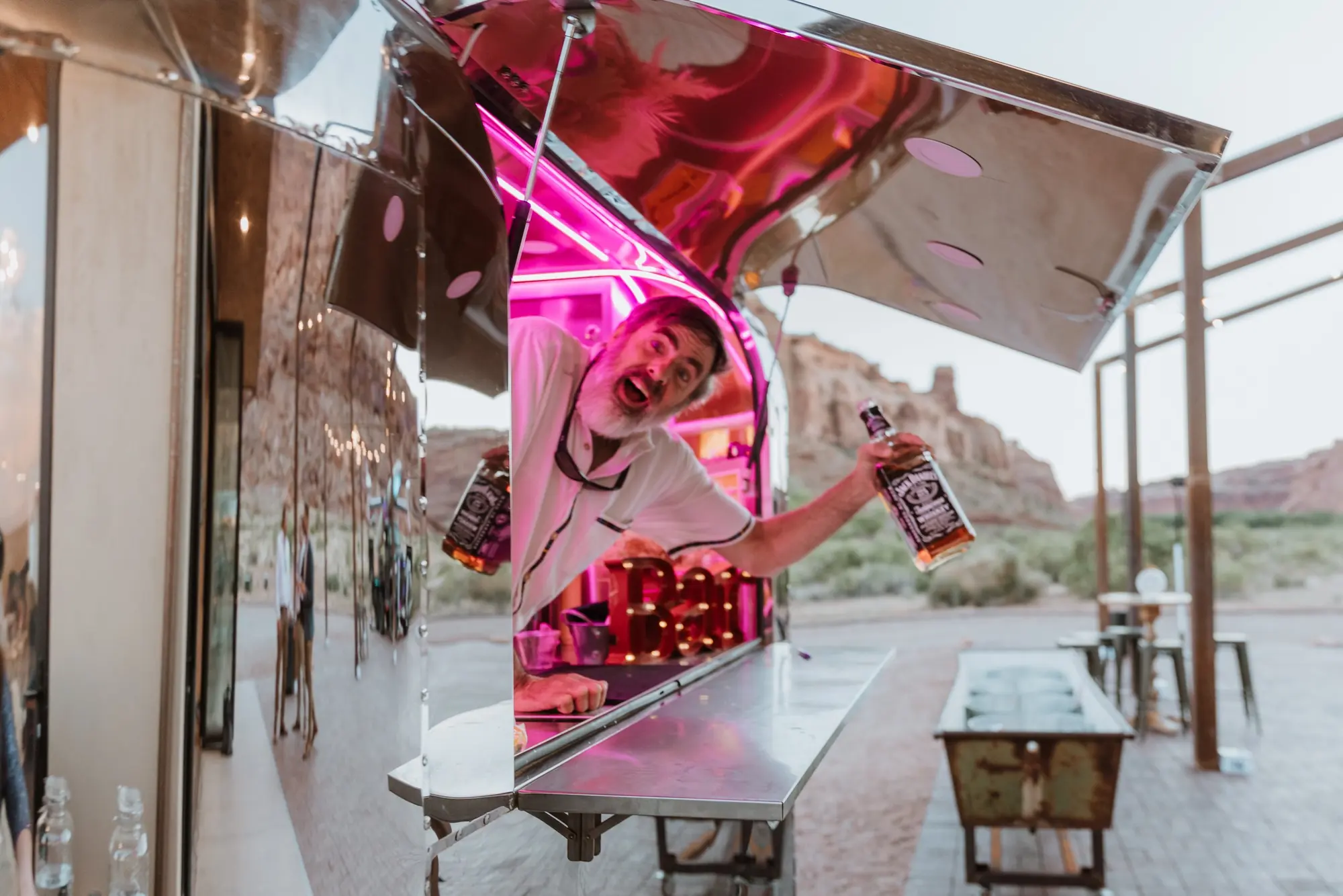 A man with a beard holding a Jack Daniel's bottle and a glass, looking out from a reflective metal food truck with pink neon lights, outdoor setting with a rocky background and glass bottles on the side.