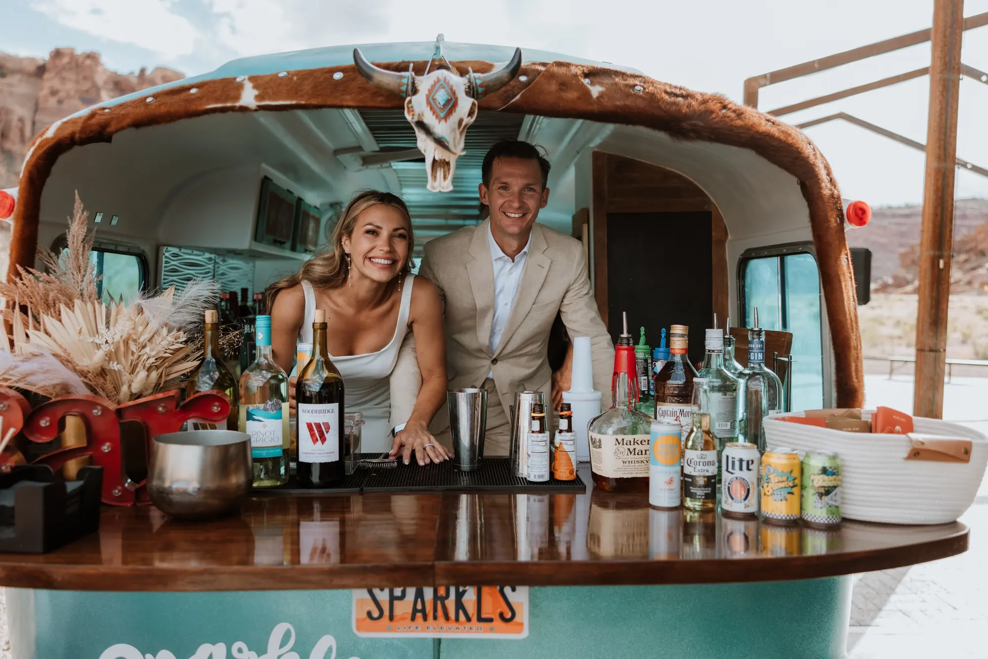 A smiling woman and man at a bar inside a vintage trailer decorated with a cow skull and animal hide, with bottles of alcohol, mixers, and bar accessories on the counter, set outdoors with a rocky landscape.