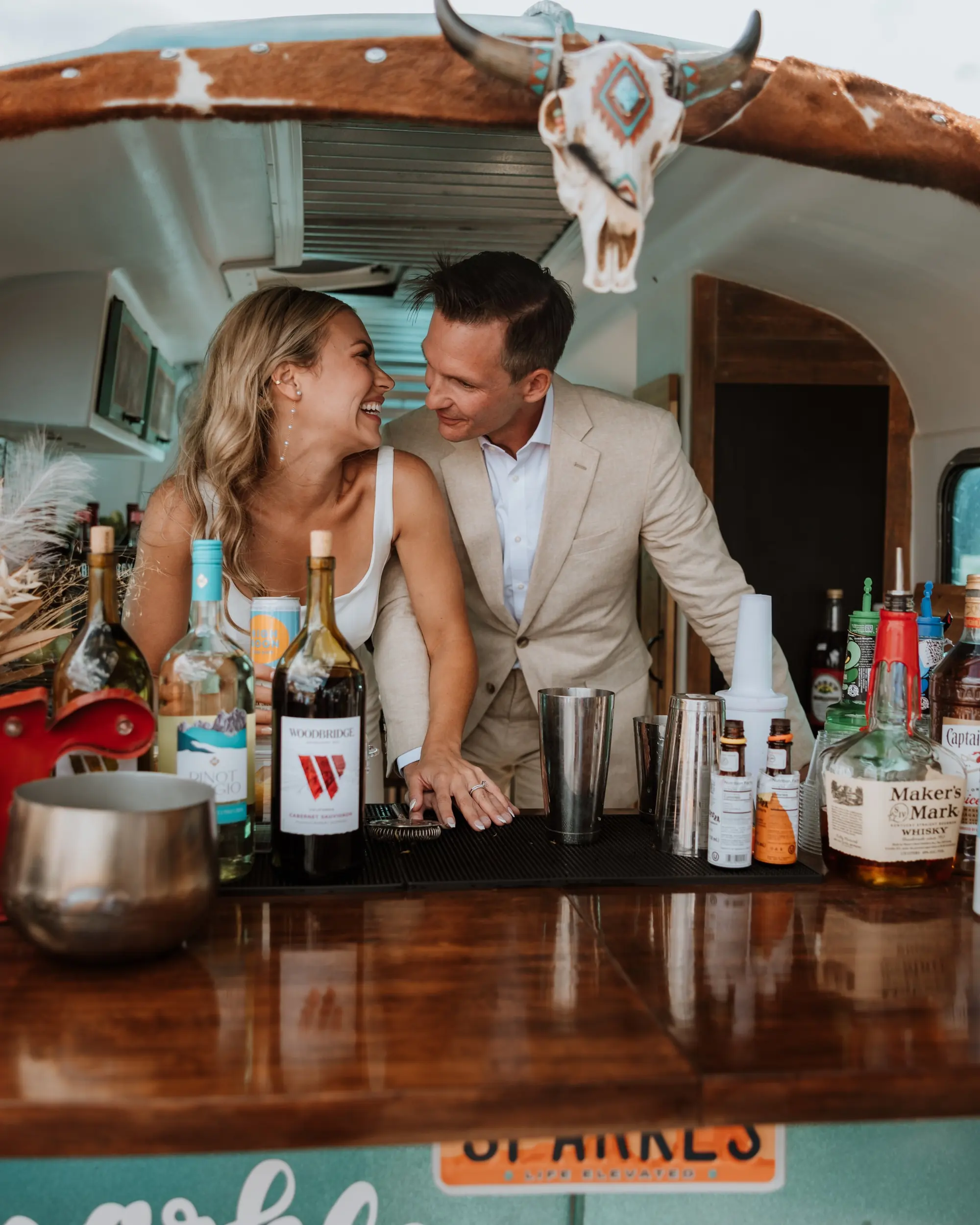 A smiling woman and man enjoy each other's company behind a bar with bottles of alcohol, mixers, and bar tools. The bar has a wood countertop and a Texas license plate is visible beneath the counter. A decorative cow skull hangs from the ceiling.