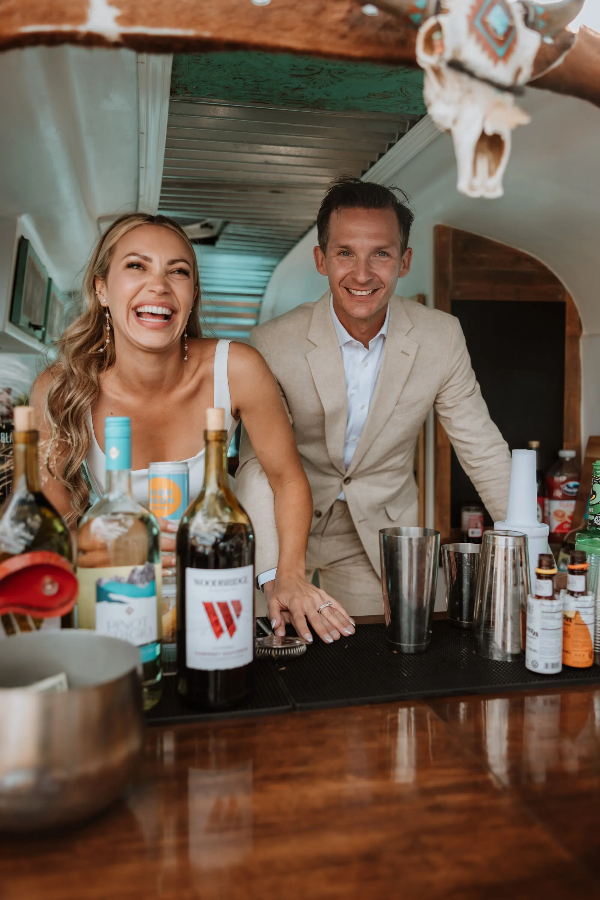 A man and woman smiling behind a bar with various bottles, shakers, and bar tools, in a cozy indoor setting with wooden accents.
