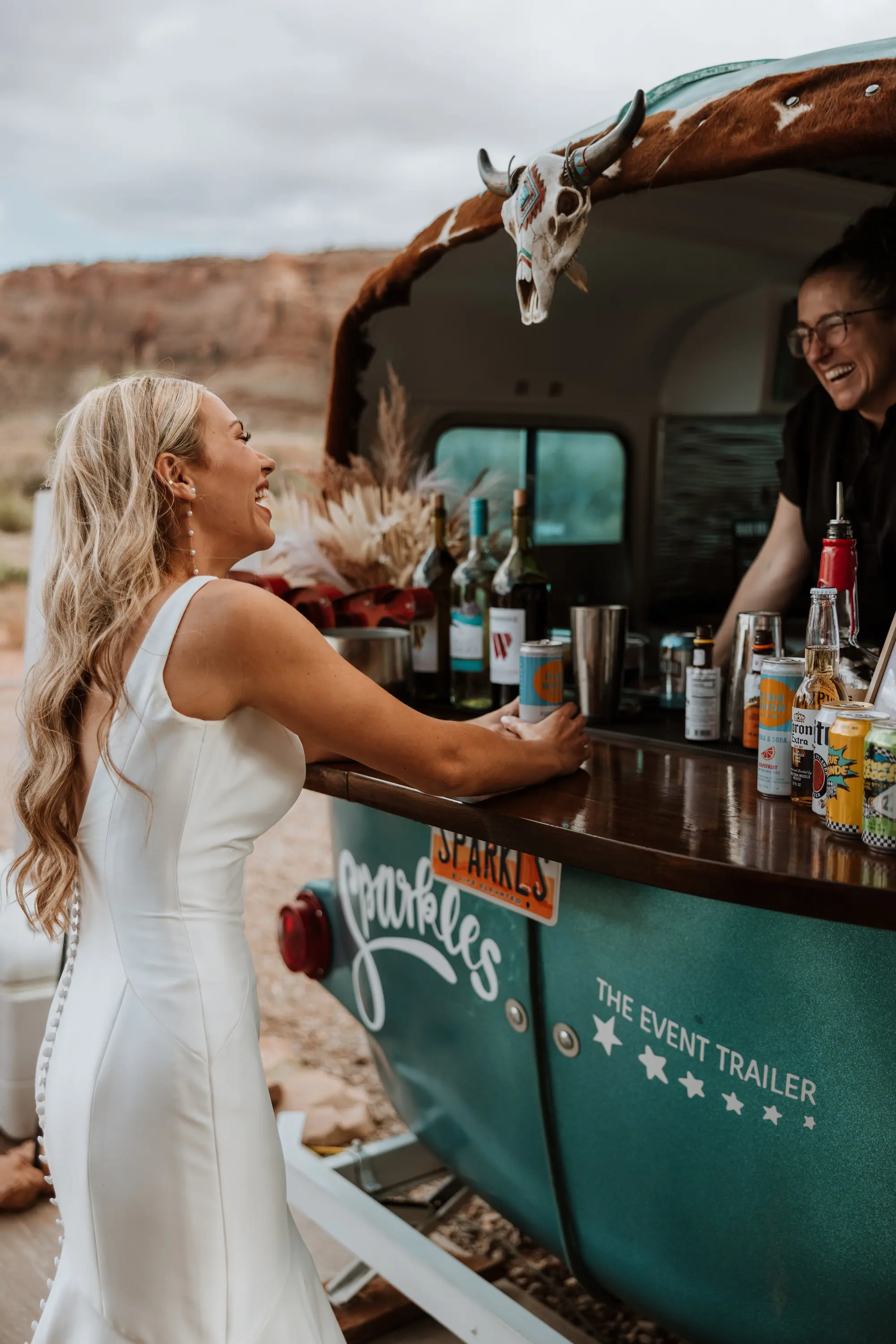 A woman in a white dress smiling and talking to a man behind a beverage trailer decorated with a cow skull and a sign that says "Sparkles". The trailer is set outdoors with a natural landscape of rocky hills in the background.