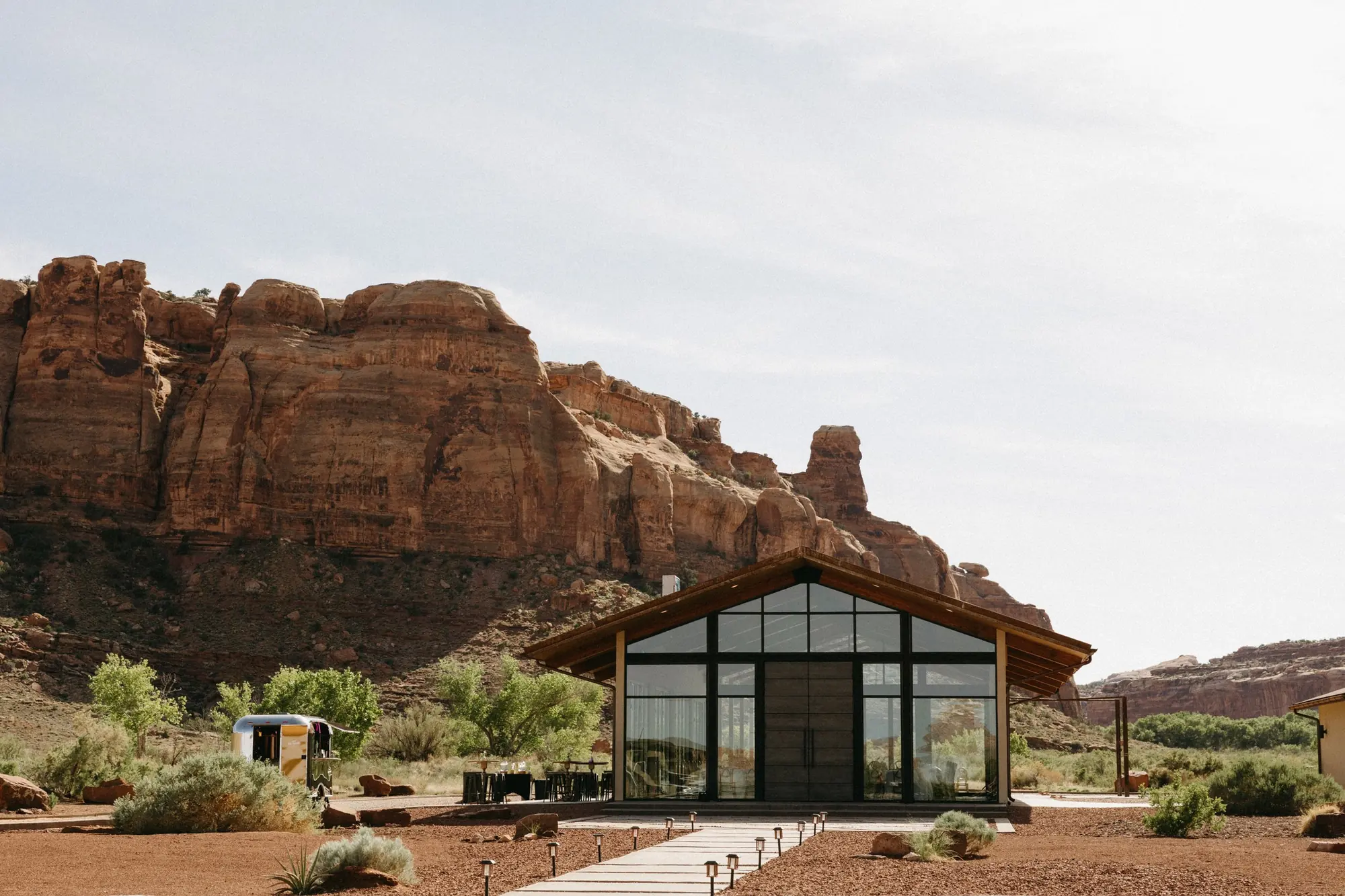 Modern glass house with pointed roof, set against red rock formations and mountain cliffs, with a pathway and sparse desert vegetation in the foreground.