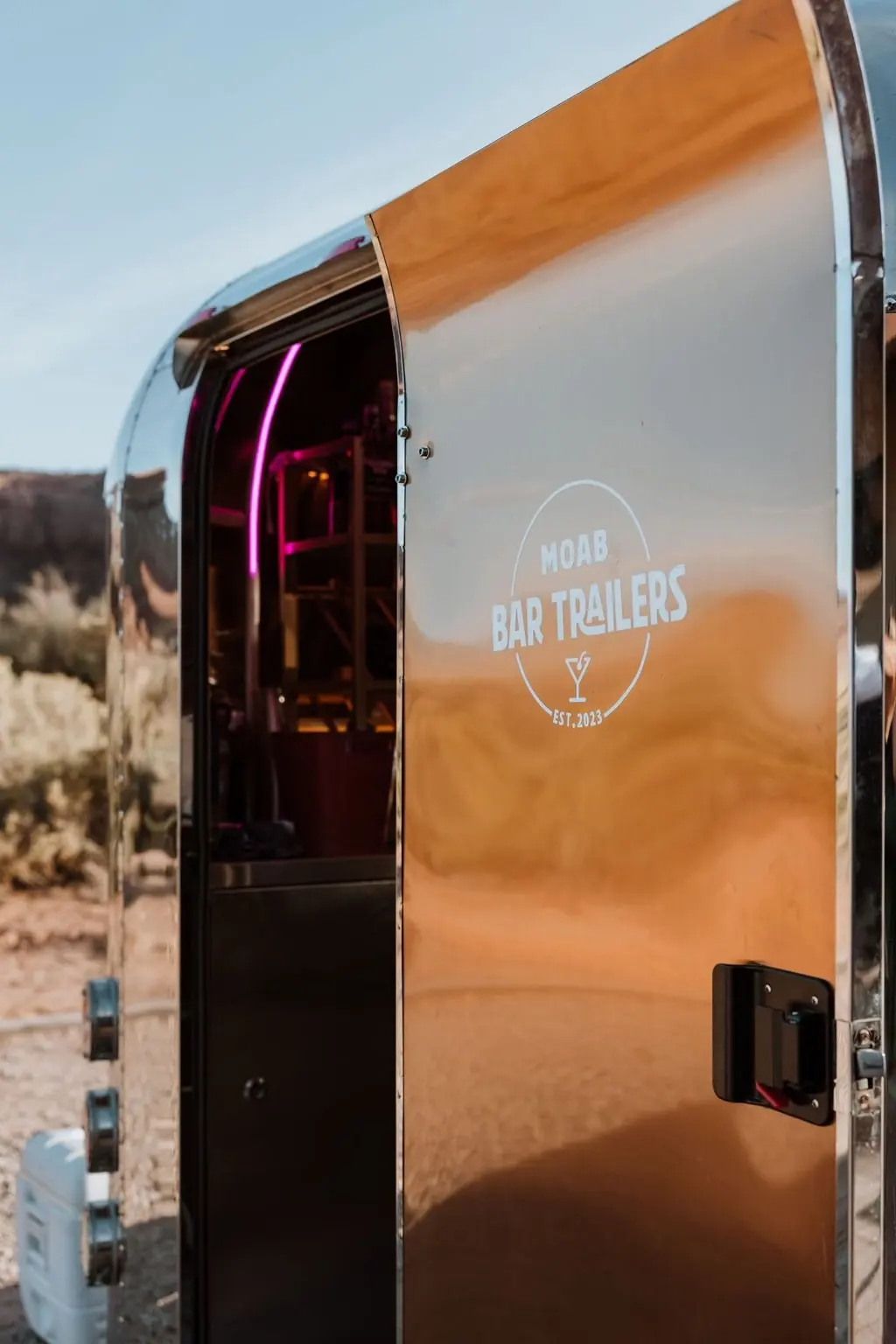 A reflective silver bar trailer with a wooden panel, displaying a logo that reads 'MOAB BAR TRAILERS EST. 2023' with a martini glass icon, situated outdoors in a desert landscape.