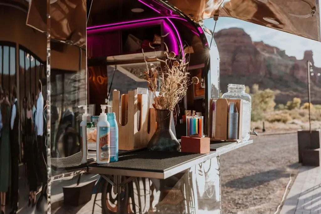 A portable coffee or snack stand set up outdoors in a desert landscape, with bottles of sanitizer, a large jar, colorful cups, and drying mats visible, and rugged terrain with hills in the background.