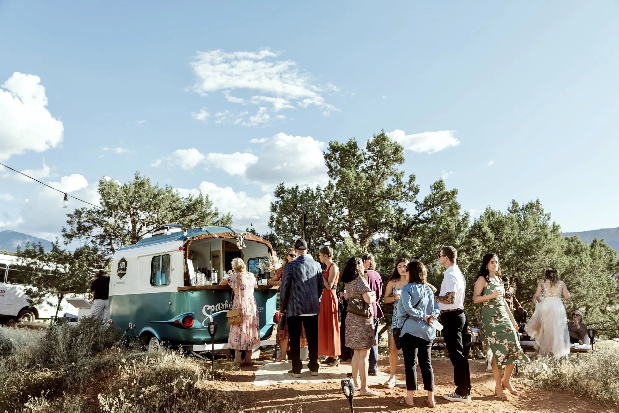 A group of people gathered outdoors around a small mobile food or beverage cart in a natural setting with trees and mountains in the background.