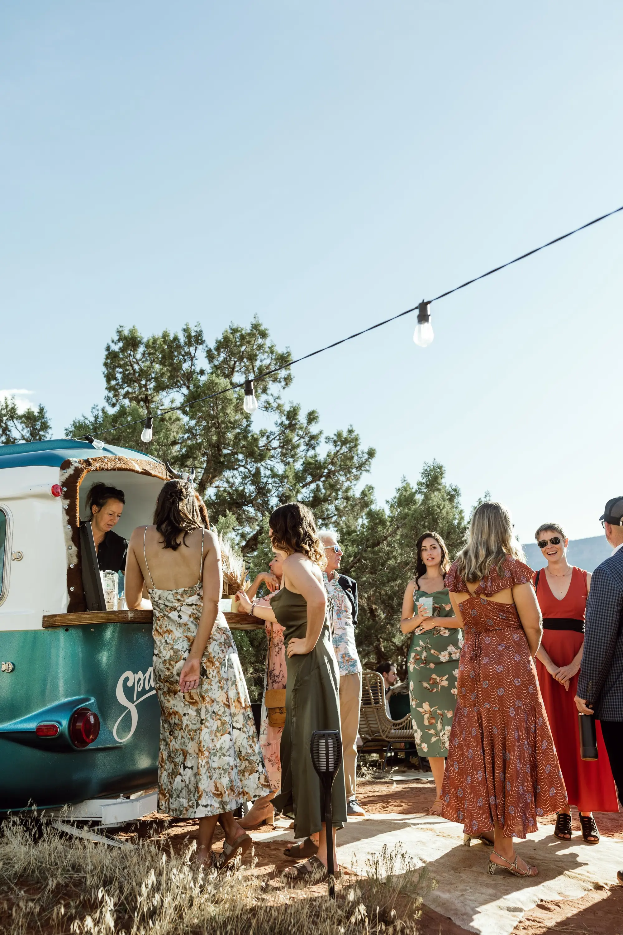 A group of women gathered outdoors around a teal ice cream truck, wearing summer dresses with trees and string lights overhead on a sunny day.