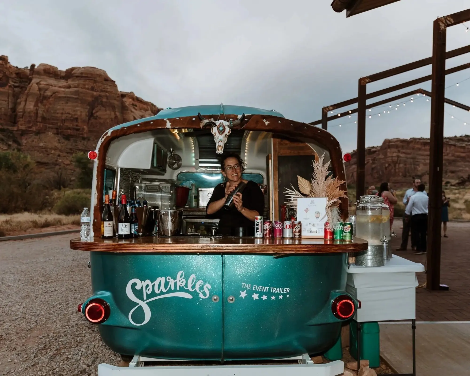 A woman standing inside a teal vintage-style trailer selling Sparkele's Coca-Cola and other beverages, with rocky desert landscape and cliffs in the background, decorated with a bull skull hanging above the window.