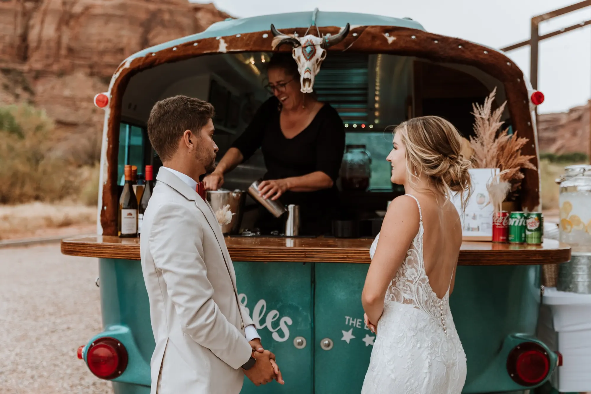 A bride and groom holding hands and smiling at each other in front of a teal food truck decorated with a cow skull and dried plants, with a desert landscape in the background.