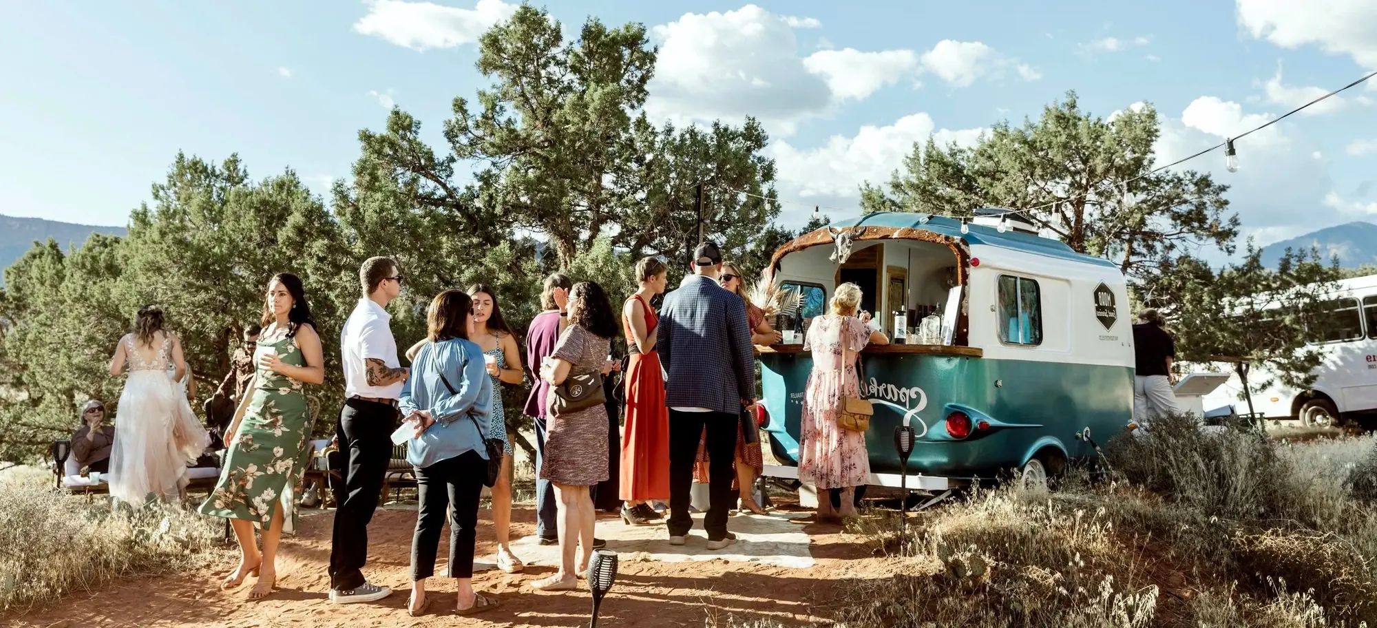 A group of people gathered around a teal and white food truck in an outdoor setting with trees and mountains in the background.