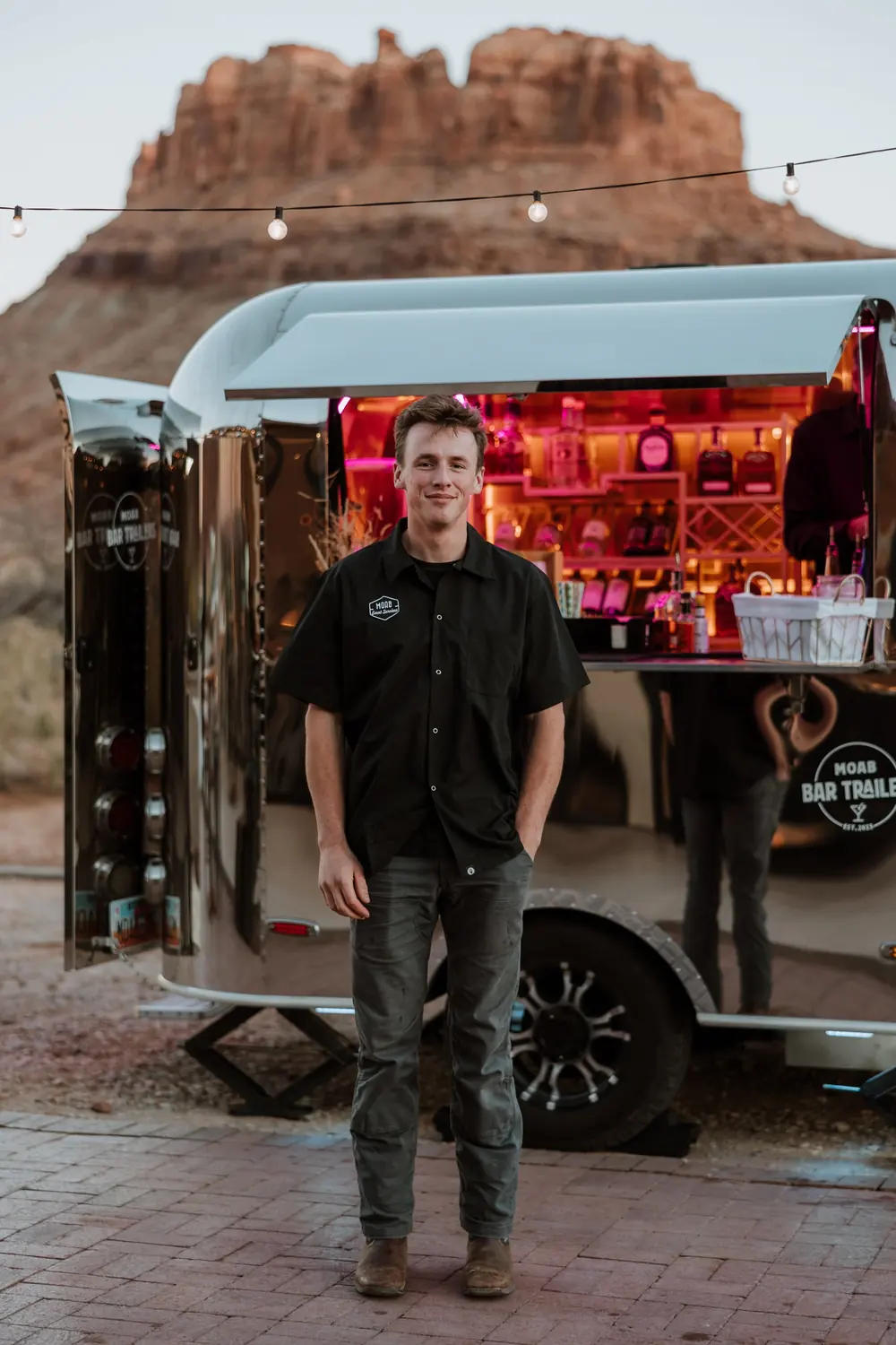 Riley, owner of Moab Bar Trailers, standing in front of a mobile bar trailer at dusk in Moab, Utah