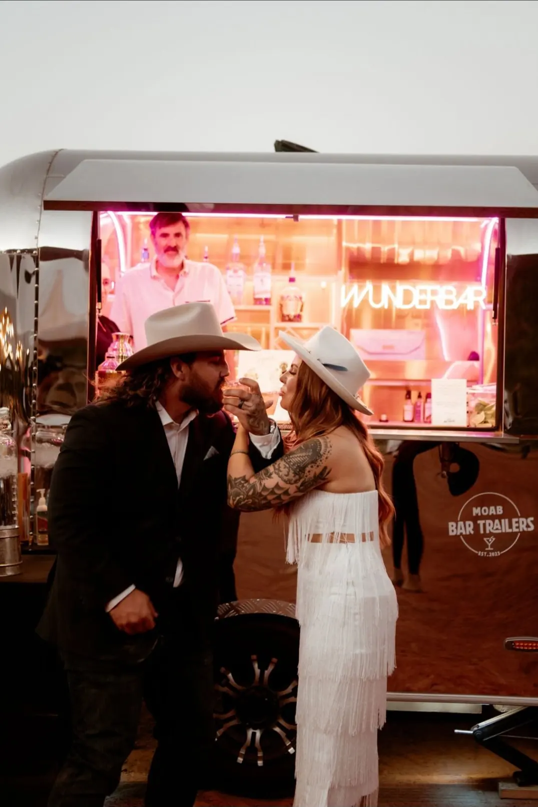 A man and woman with tattoos and wearing cowboy hats sharing a drink in front of a neon-lit bar trailer named WunderBar, with a bartender inside.