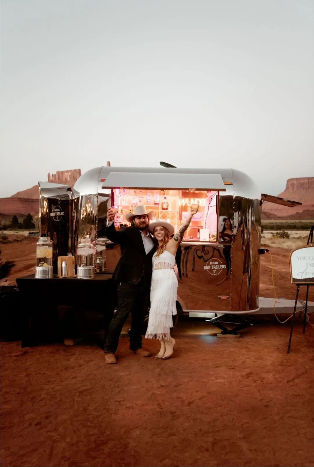 A couple dressed in Western attire standing in front of a shiny silver concession trailer in a desert landscape, they are smiling and raising their glasses, with a woman waving. The trailer has a neon sign and bar items nearby.