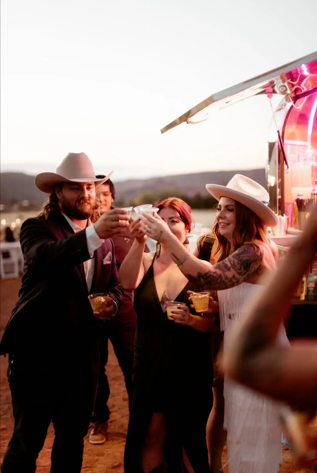 A group of friends wearing cowboy hats clink glasses together at an outdoor evening gathering near a colorful food truck.