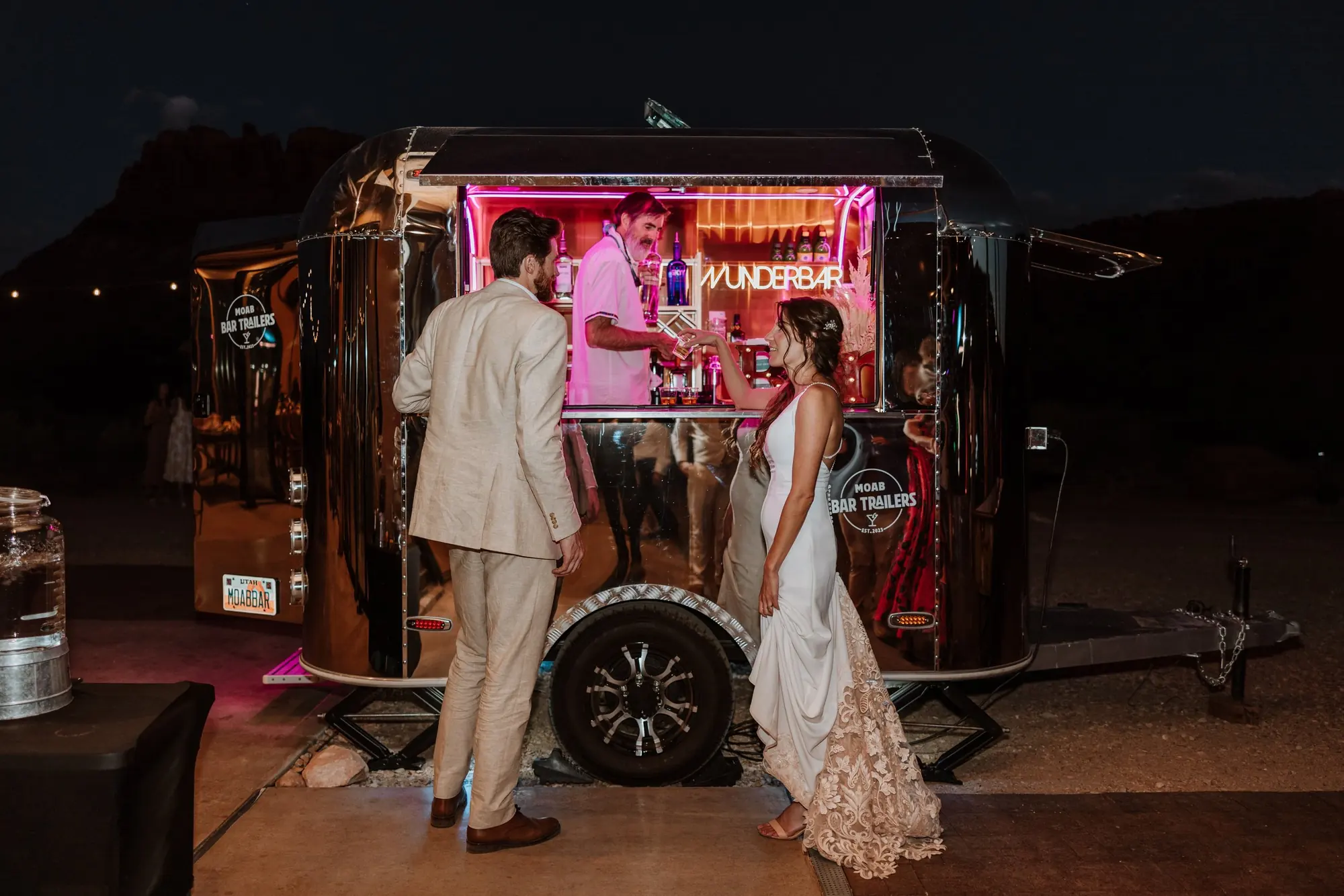 A nighttime scene with a reflective silver mobile bar trailer labeled 'Munderbar' serving drinks. A man in a beige suit and a woman in a white dress are sharing a moment near the bar, with the woman touching the man's arm. The bartender, behind the b