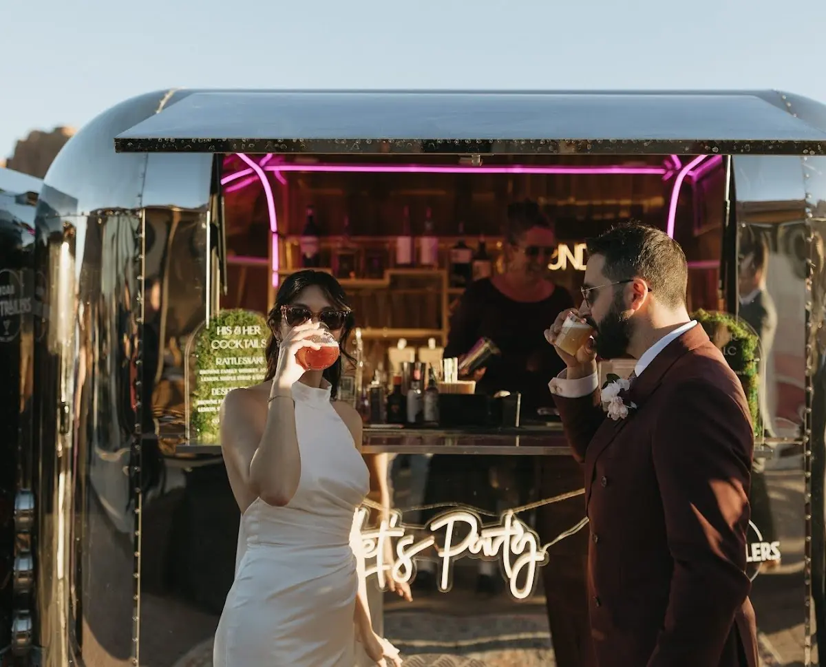 A man and woman in formal attire enjoying drinks at a mobile bar with neon lighting, outdoors during sunset.