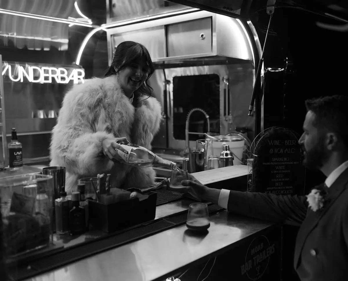 A woman with a fur coat serving drinks to a man in a suit at a bar inside a trailer.