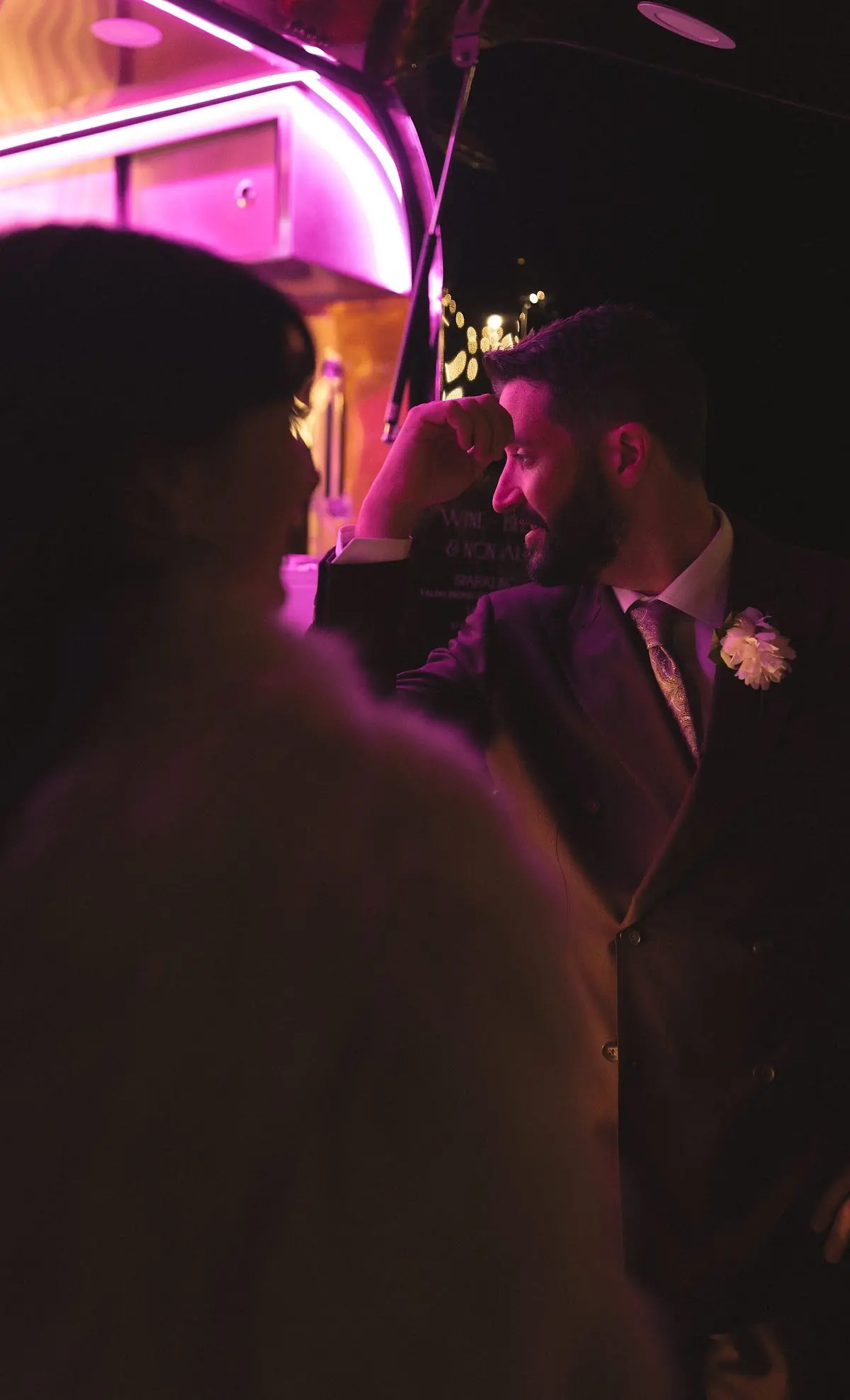 A man in formal attire with a white flower on lapel, illuminated by pink and purple lighting, smiling and resting his head on his hand at a social event or party.