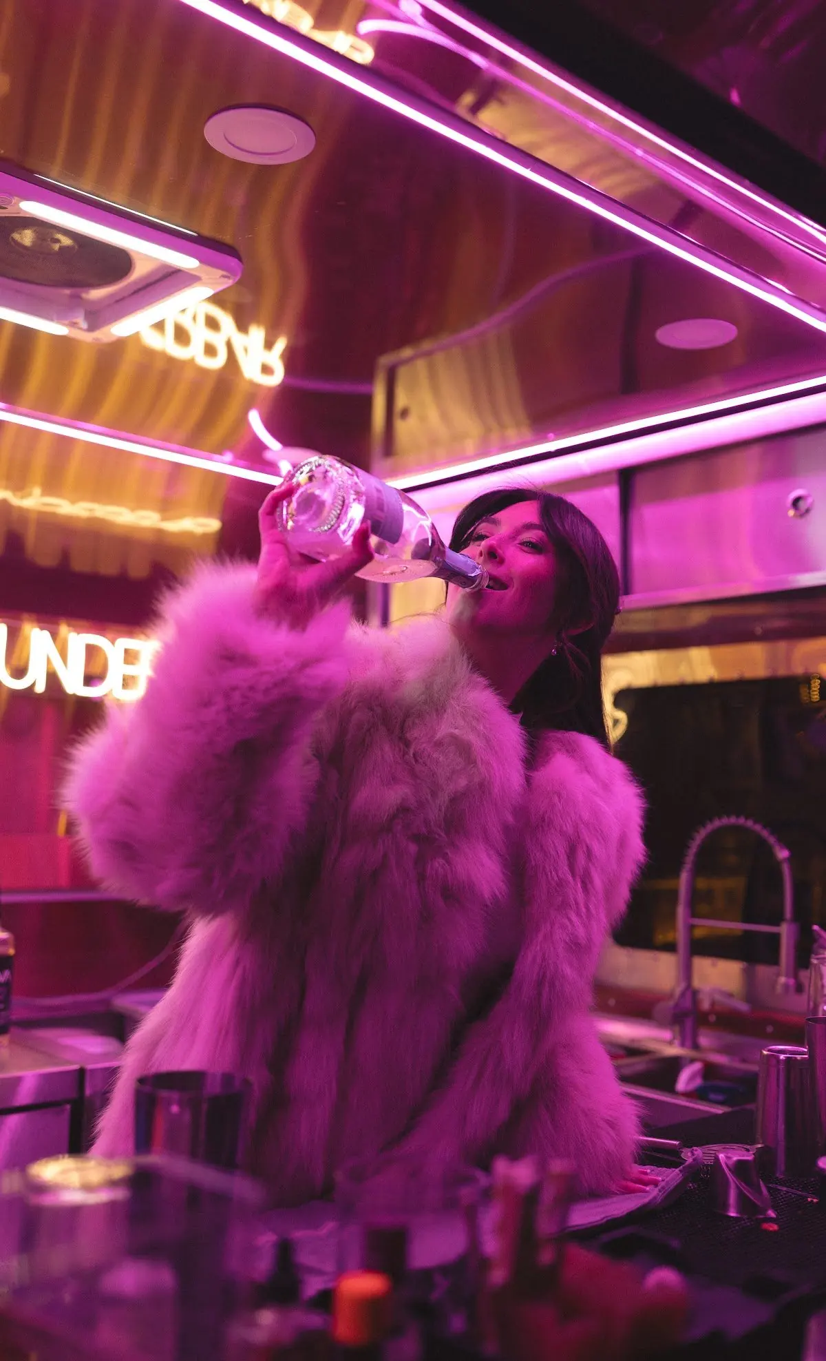 A woman wearing a fluffy pink coat is drinking from a bottle in a neon-lit bar or club with pink and purple lighting.
